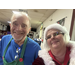 A man in a green apron stands smiling next to Mrs. Claus